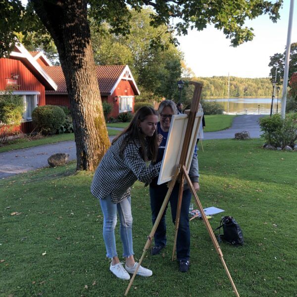 Två stycken står och målar på en tavla på ett stativ.