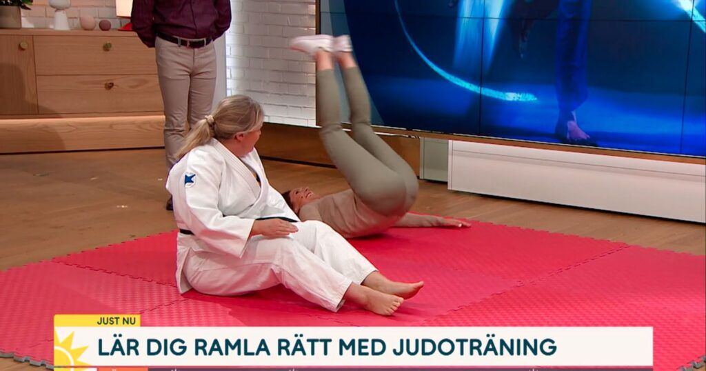 Judoinstruktör sitter på judomatta i studion på TV4. Bredvid rullar programledaren bakåt på rygg för att öva bakåtfall.