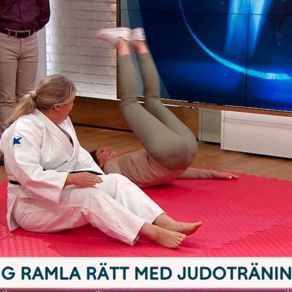 Judoinstruktör sitter på judomatta i studion på TV4. Bredvid rullar programledaren bakåt på rygg för att öva bakåtfall.