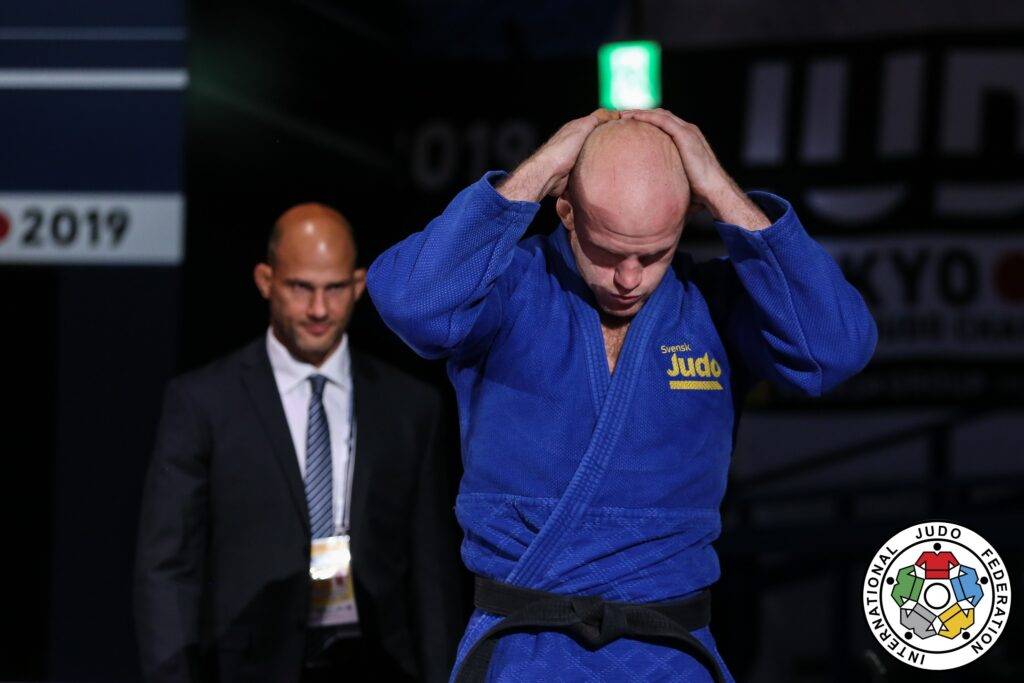 Marcus Nyman i blå judodräkt går in på arenan. Tittar ner och håller båda händer knäppta bakom huvudet. Landslagschef Robert Eriksson går bakom.