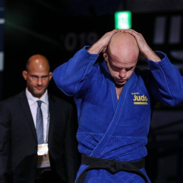 Marcus Nyman i blå judodräkt går in på arenan. Tittar ner och håller båda händer knäppta bakom huvudet. Landslagschef Robert Eriksson går bakom.