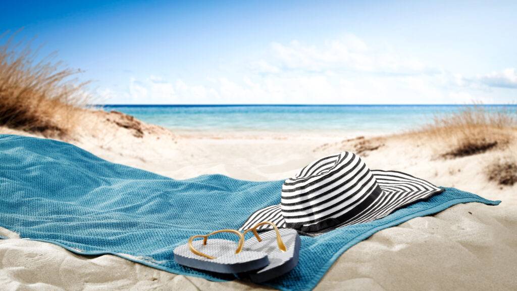 Randig hatt och flip-flops på en blå handdduk på en strand.