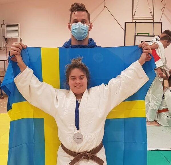 Amanda Orrbo i vit judodräkt med silvermedalj runt halsen. Bakom henne svenska flaggan och tränaren Jonathan Fagerhill.