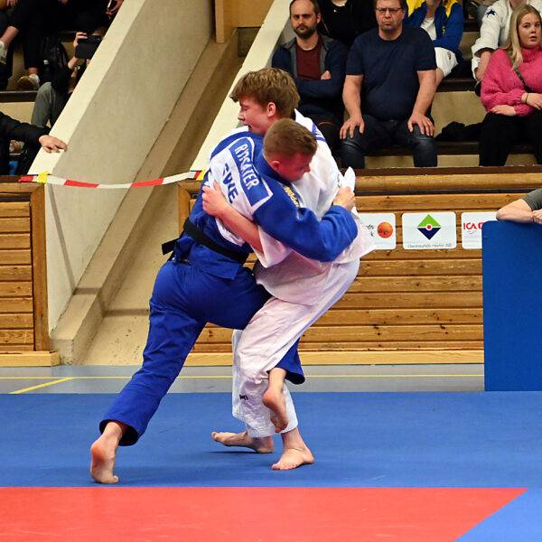 Två killar i judodräkt står på judomattan. Den ena har krokat sitt ben runt den andra och försöker fälla honom.